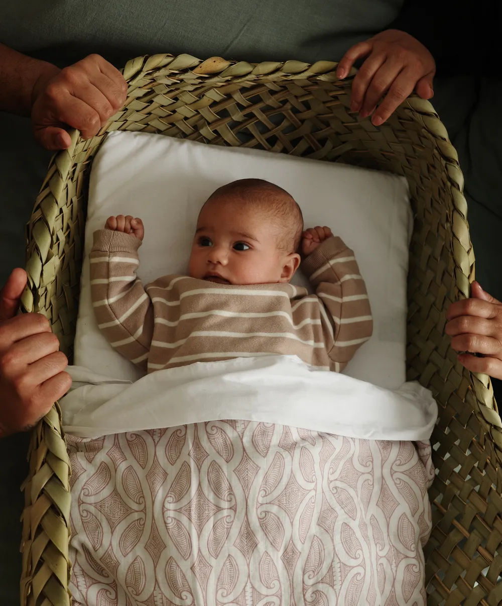 Whānau hands cradling a pēpi resting on their back in a wahakura
